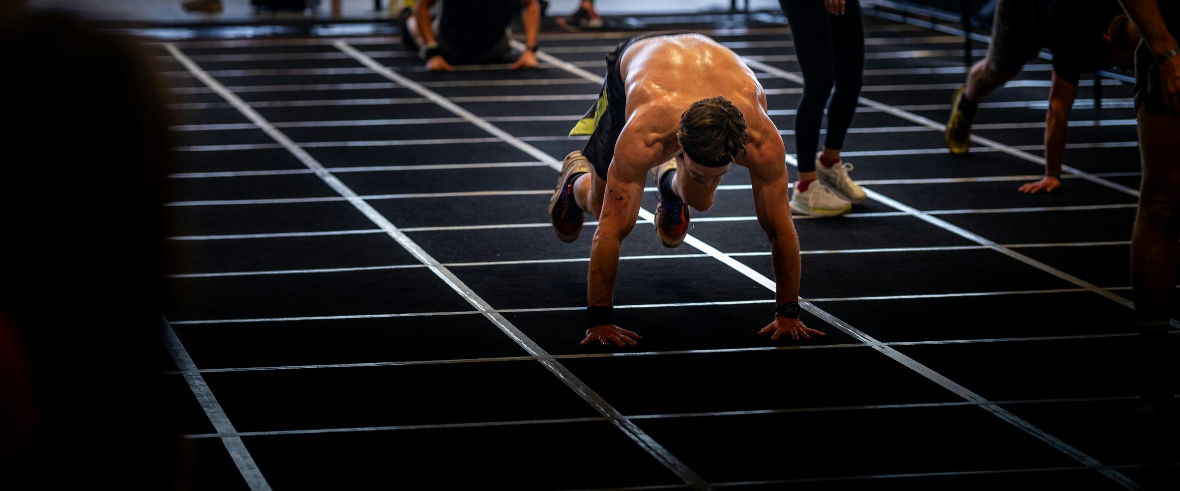 A burpee broad jump down a marked lane in a dark arena, other people training behind